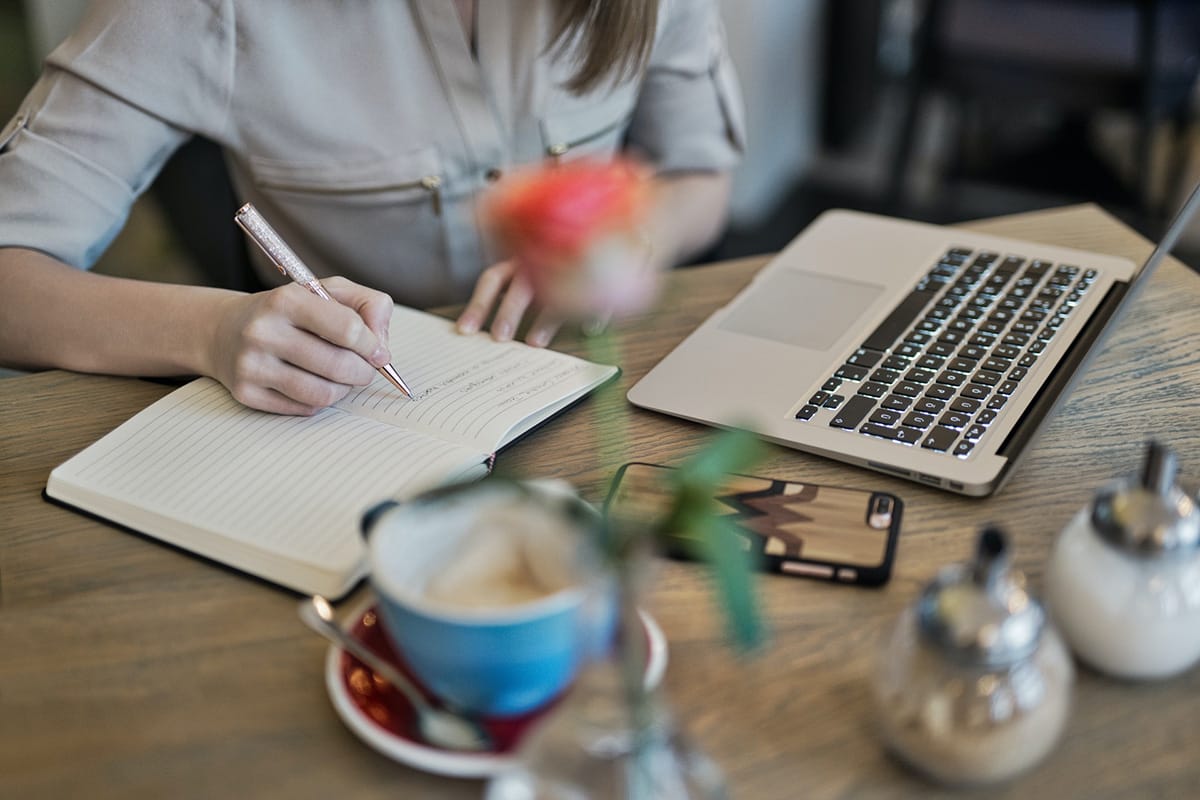 Person writing in a notebook next to an open laptop, smartphone, and coffee cup on a wooden table.