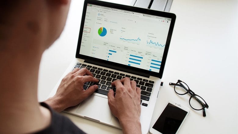 A person is working on a laptop displaying graphs and charts, with a smartphone and a pair of eyeglasses placed on the table beside them.