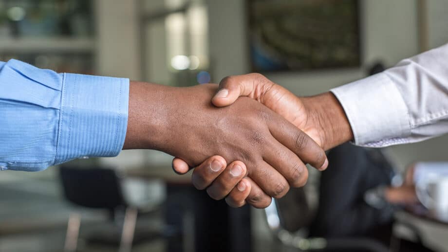Two individuals shaking hands in an office setting, one wearing a blue shirt and the other in a white shirt, indicating a professional greeting or agreement.
