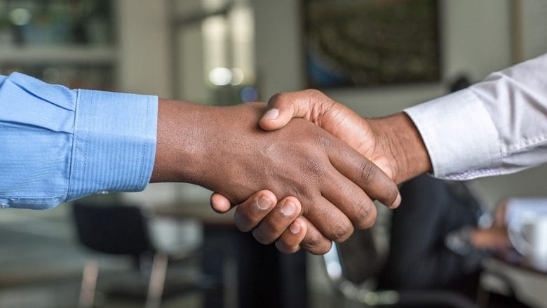 Two individuals shaking hands in an office setting, one wearing a blue shirt and the other in a white shirt, indicating a professional greeting or agreement.