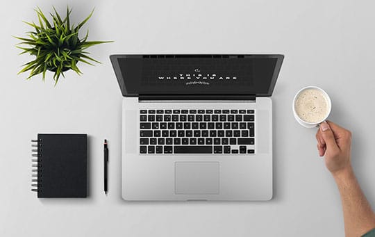 Top view of a workspace with a notebook, pen, laptop, and a hand holding a cup of coffee next to a small plant on a white surface.