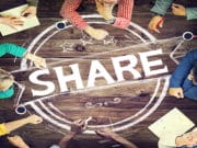 A group of people sit around a wooden table with the word SHARE and circular graphics drawn in white on the tabletop.