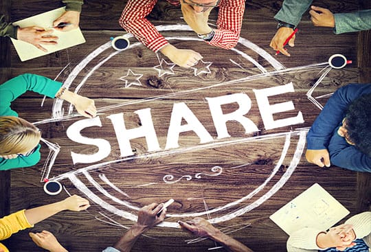 A group of people sit around a wooden table with the word SHARE and circular graphics drawn in white on the tabletop.