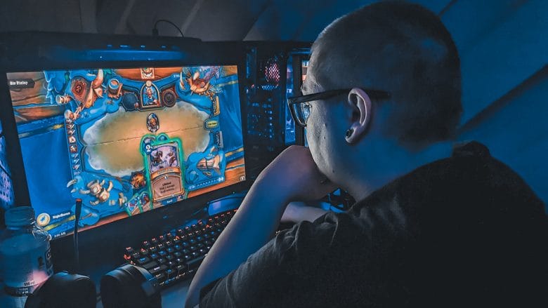 A person is intently playing a digital card game on a computer with a large monitor in a dimly lit room.