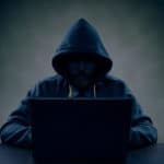 A person wearing a hoodie sits in front of a laptop in a dimly lit setting.