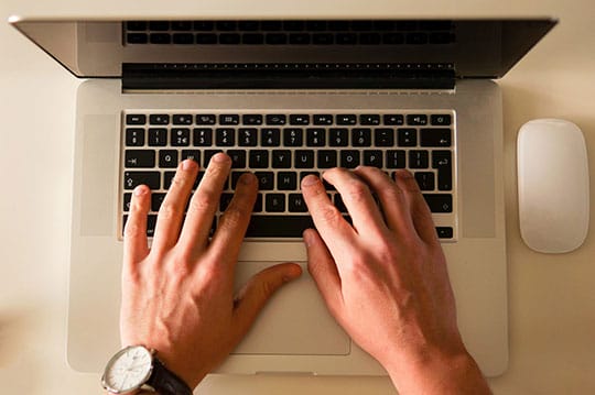 A person types on a laptop keyboard, with a smartwatch on their left wrist and a wireless mouse beside the computer.
