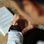 A person interacts with a smartwatch on their wrist, with a book or document featuring highlighted text in the background on a table.