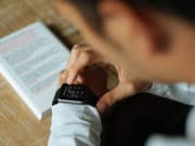 A person interacts with a smartwatch on their wrist, with a book or document featuring highlighted text in the background on a table.