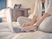 Person sitting cross-legged on a bed, typing on a laptop.
