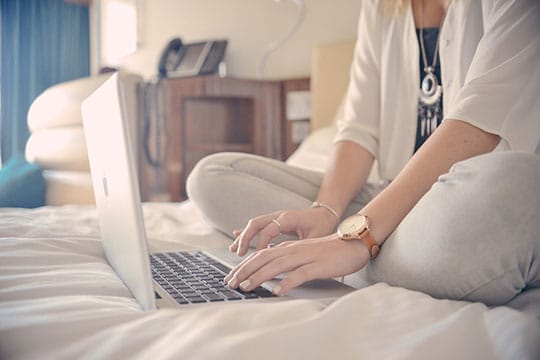 Person sitting cross-legged on a bed, typing on a laptop.
