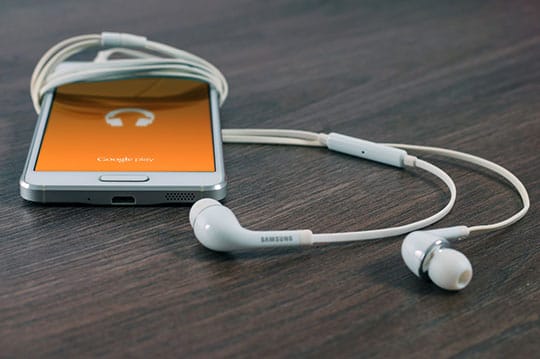 Smartphone with an orange display showing a headphone icon is placed on a wooden surface, accompanied by white Samsung earphones.
