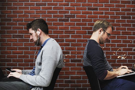 Two men sit back-to-back against a brick wall, using laptops; one is wearing earphones.