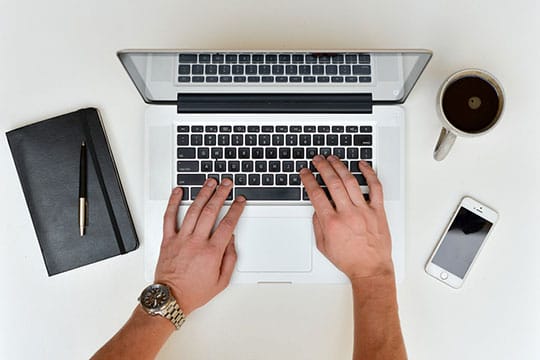 A person types on a laptop at a desk with a notebook, pen, coffee cup, and smartphone.