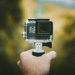 A hand holding a small action camera mounted on a handheld grip, with a blurred outdoor background.