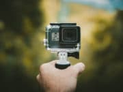 A hand holding a small action camera mounted on a handheld grip, with a blurred outdoor background.