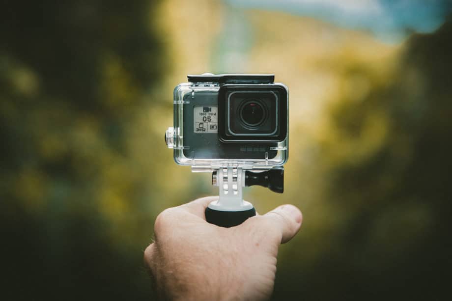 A hand holding a small action camera mounted on a handheld grip, with a blurred outdoor background.