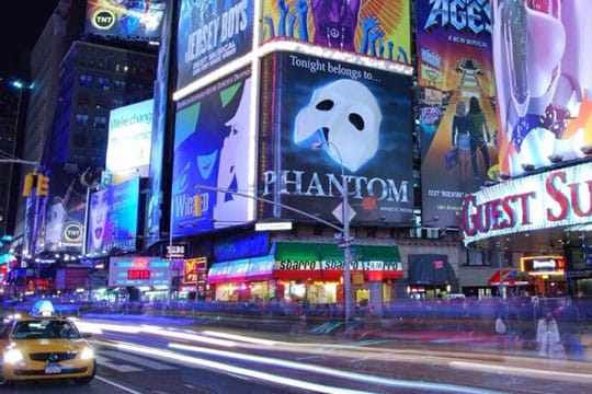 Times Square at night, featuring illuminated billboards and advertisements, including one for "Phantom." Light trails from moving cars are visible on the street below.
