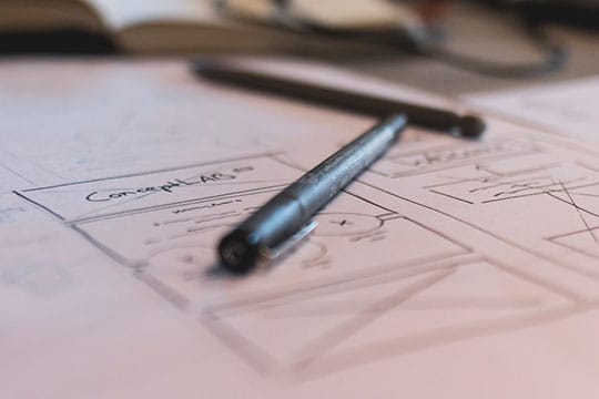 A black pen rests on sheets of paper with hand-drawn diagrams and notes; a blurred notebook is in the background.