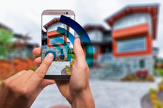 Person holding a smartphone in front of a modern house, capturing it with a Wi-Fi symbol overlay on the screen.