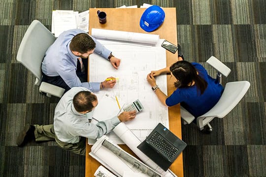 Three people reviewing architectural plans at a table with a laptop, calculator, and blue hard hat. Top-down view.