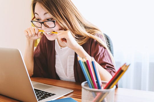 Woman wearing glasses bites a pencil while looking at a laptop screen, sitting at a desk with colored pencils in the foreground.