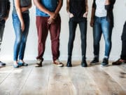 Six people stand side by side against a white wall, shown from shoulders down, wearing casual clothing and different styles of shoes.