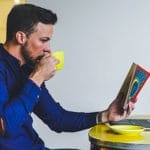 A man sits at a table, drinking from a yellow cup while reading a book and working on a laptop.