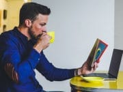 A man sits at a table, drinking from a yellow cup while reading a book and working on a laptop.