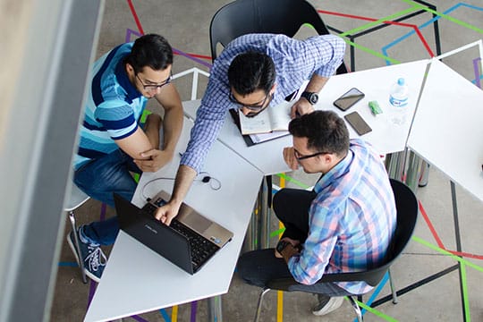 Three people sit around a table, looking at a laptop and taking notes, with phones and a water bottle nearby.