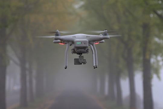 A drone with a mounted camera hovers in the middle of a tree-lined road on a foggy day.