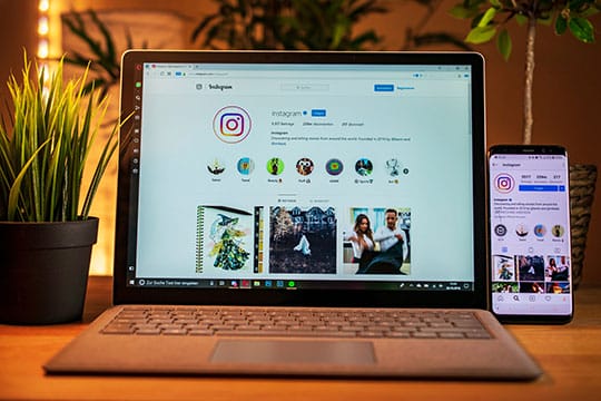A laptop and a smartphone display an Instagram profile page on a wooden desk with potted plants.