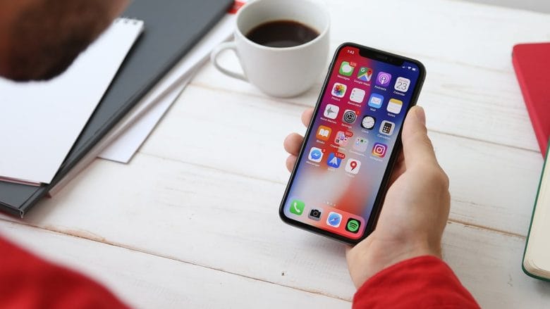 Person holding a smartphone displaying multiple app icons, with a coffee cup and notebooks on a table.