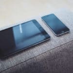 A gray couch with a tablet and phone on it.