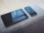 A gray couch with a tablet and phone on it.