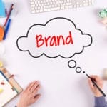 A person draws a thought bubble with the word Brand on a white desk surrounded by office and art supplies.