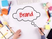 A person draws a thought bubble with the word Brand on a white desk surrounded by office and art supplies.