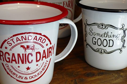 Two vintage-style mugs on a wooden surface. One mug has "Organic Dairy" branding, and the other reads "Something Good.