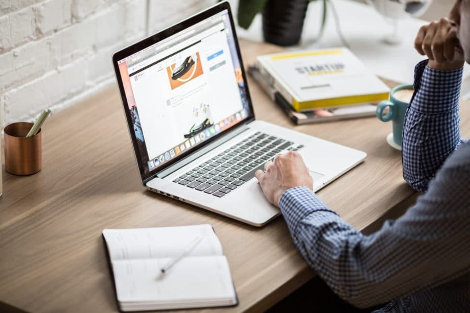 Person using a laptop on a wooden desk with a notebook, coffee mug, and books nearby. A sneaker is displayed on the laptop screen.