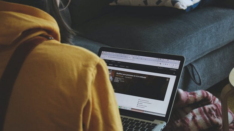 Person in a yellow hoodie using a laptop on a couch with a colorful pillow, screen showing a website.