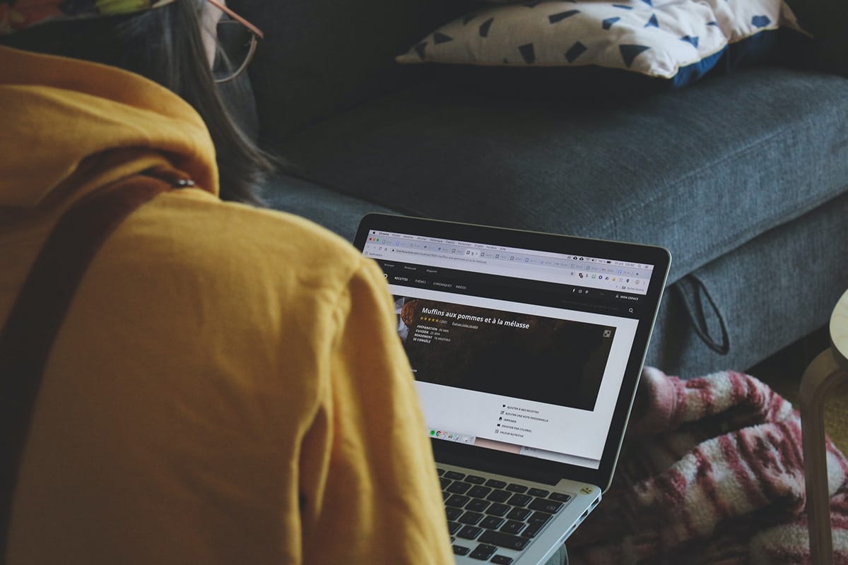 Person in a yellow hoodie using a laptop on a couch with a colorful pillow, screen showing a website.