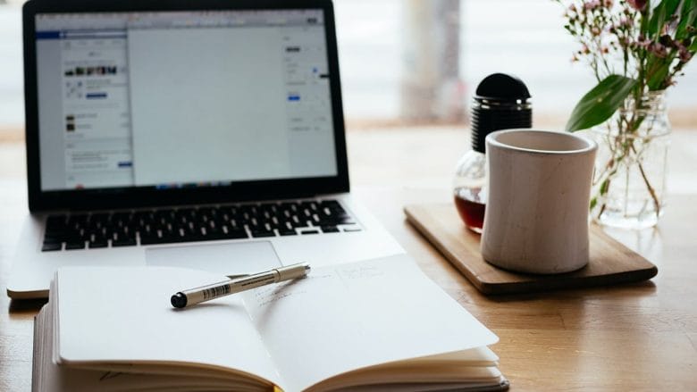 Open laptop, notebook with pen, coffee mug, and flowers on a wooden desk, suggesting a workspace or study area.