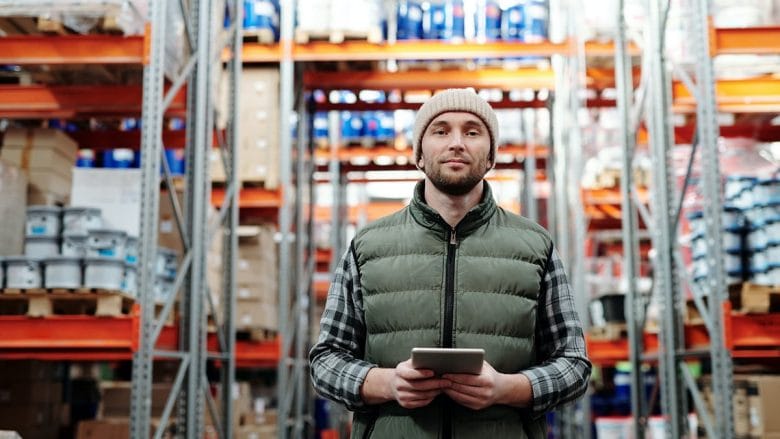 A person wearing a beanie and vest holds a tablet while standing in a warehouse with shelves stocked with various items.
