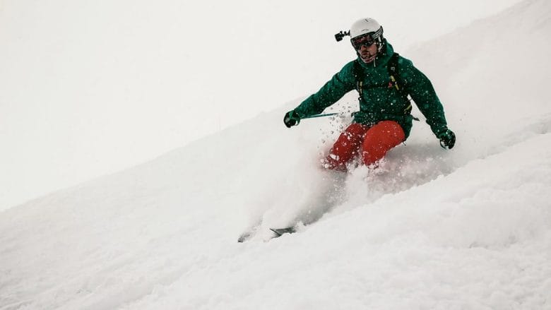 A person wearing a helmet and goggles skis downhill through deep snow, creating a spray of powder around them.