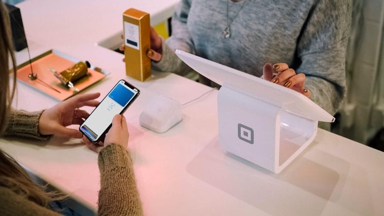 A person holds a phone to make a contactless payment at a white Square register while another person stands behind the counter.