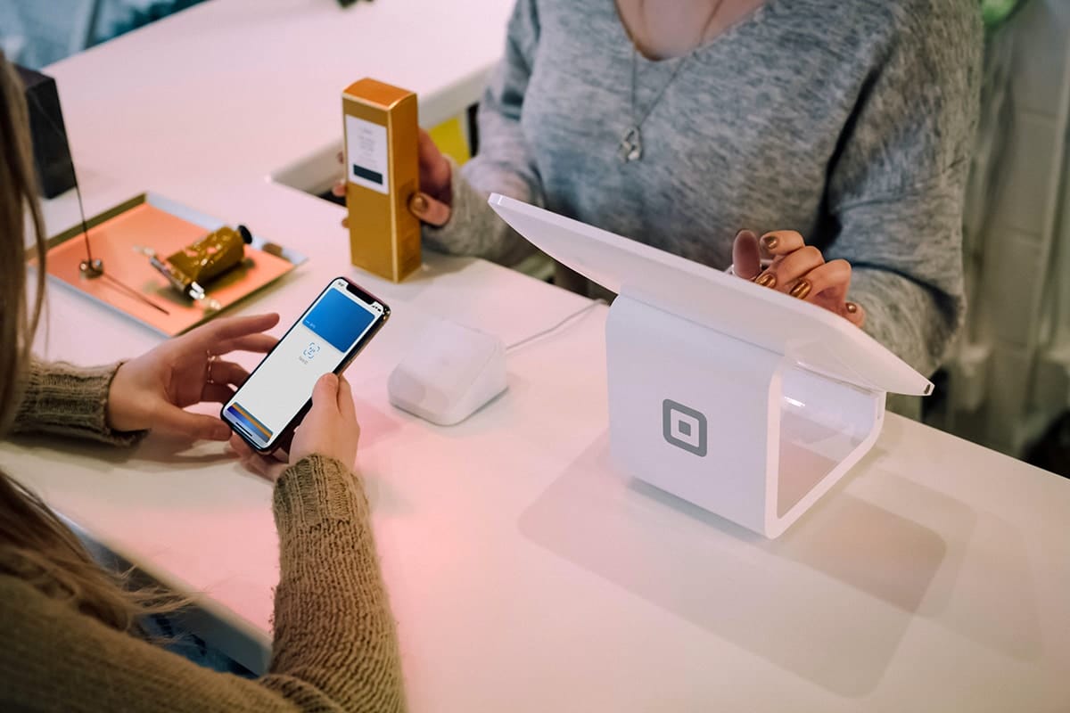 A person holds a phone to make a contactless payment at a white Square register while another person stands behind the counter.