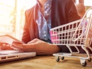 A person is holding a smartphone while sitting at a table with a laptop. A small toy shopping cart is placed on the table next to the laptop. The person is wearing a dark blazer over a blue shirt. The scene is brightly lit with a soft focus background.