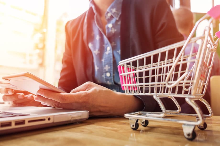 A person is holding a smartphone while sitting at a table with a laptop. A small toy shopping cart is placed on the table next to the laptop. The person is wearing a dark blazer over a blue shirt. The scene is brightly lit with a soft focus background.