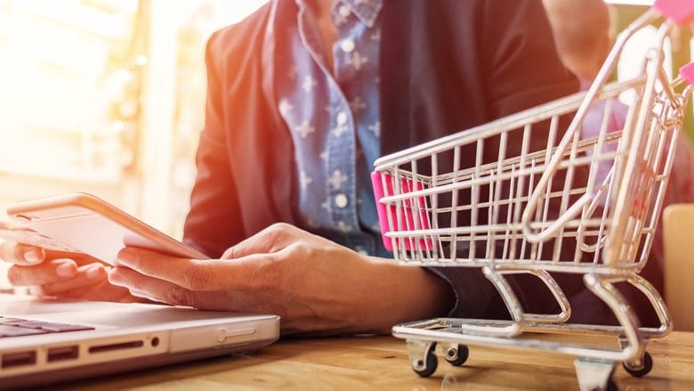 A person is holding a smartphone while sitting at a table with a laptop. A small toy shopping cart is placed on the table next to the laptop. The person is wearing a dark blazer over a blue shirt. The scene is brightly lit with a soft focus background.