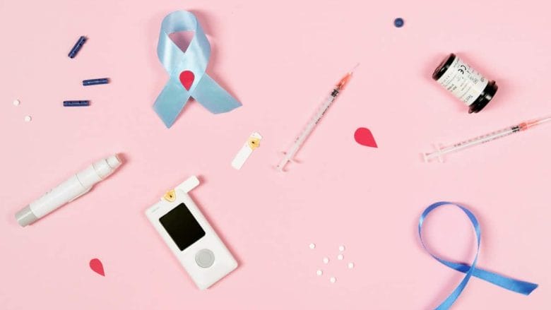 A flat lay of diabetes management tools on a pink background, including a glucose meter, test strips, lancet devices, insulin syringes, and a vial. Blue awareness ribbons are also present.