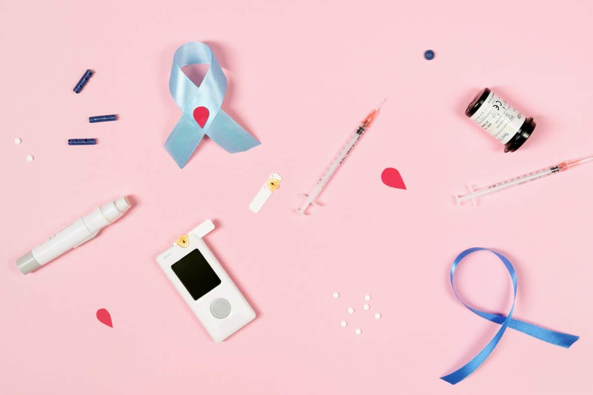 A flat lay of diabetes management tools on a pink background, including a glucose meter, test strips, lancet devices, insulin syringes, and a vial. Blue awareness ribbons are also present.
